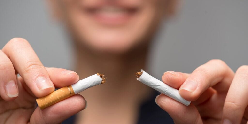 Close up portrait of young attractive woman breaking down cigarette to pieces. Studio shot selective focus isolated on grey. Addiction concept